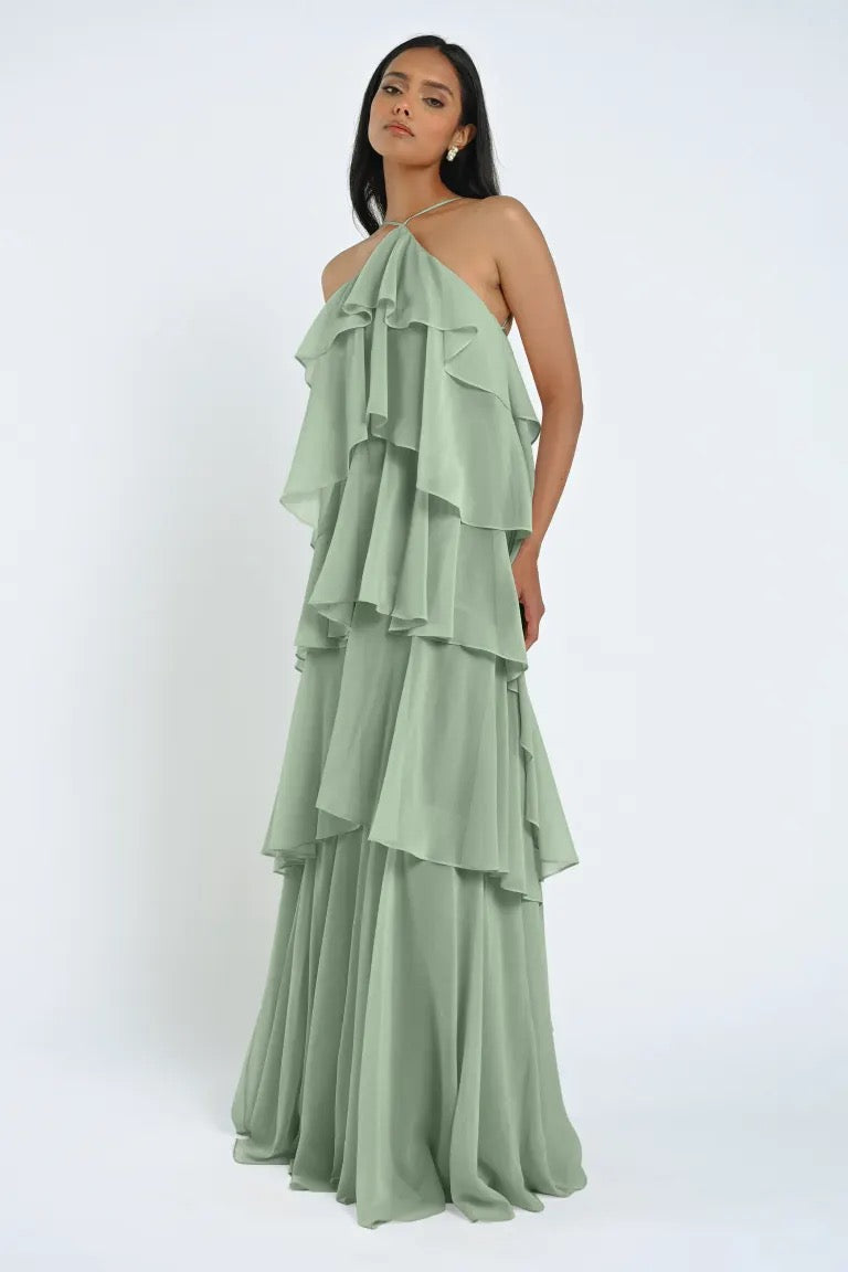 A woman wearing a light green, layered "Farley Bridesmaid Dress" by Jenny Yoo, available at Bergamot Bridal, stands against a plain white background.