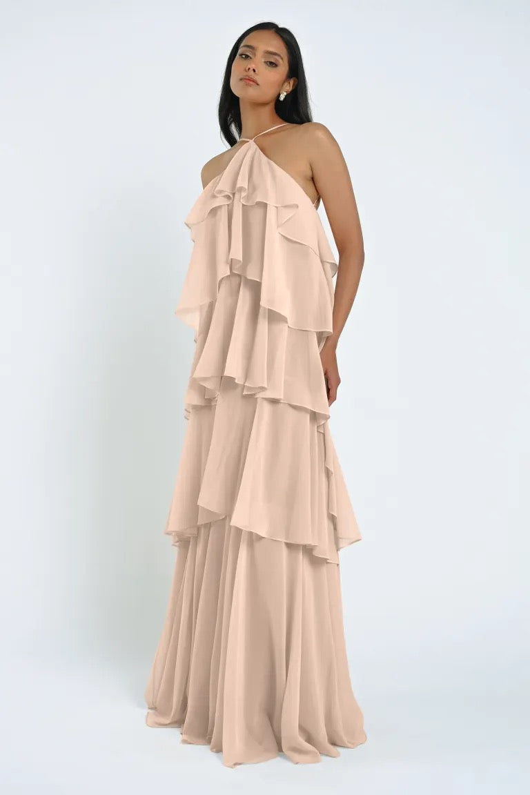 A woman stands wearing the Farley Bridesmaid Dress by Jenny Yoo from Bergamot Bridal—a long, flowy, beige off-shoulder dress featuring layered ruffles. The sheer chiffon fabric adds an ethereal touch. She has a neutral expression and her arms are relaxed at her sides.