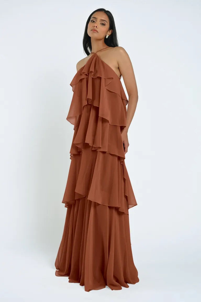 A woman in a tiered, rust-colored Farley Bridesmaid Dress by Jenny Yoo from Bergamot Bridal stands against a plain background.