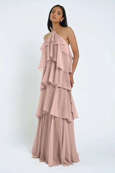 A woman stands against a plain background wearing an exquisite event dress: the sleeveless, tiered ruffle, floor-length Farley Bridesmaid Dress by Jenny Yoo in light pink sheer chiffon from Bergamot Bridal.