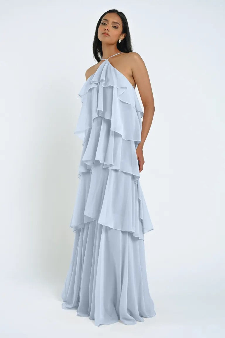 A woman stands wearing the Farley Bridesmaid Dress by Jenny Yoo, a light blue, tiered halter neck gown from Bergamot Bridal with a flowing chiffon skirt that exudes an effortless feel against a plain background.