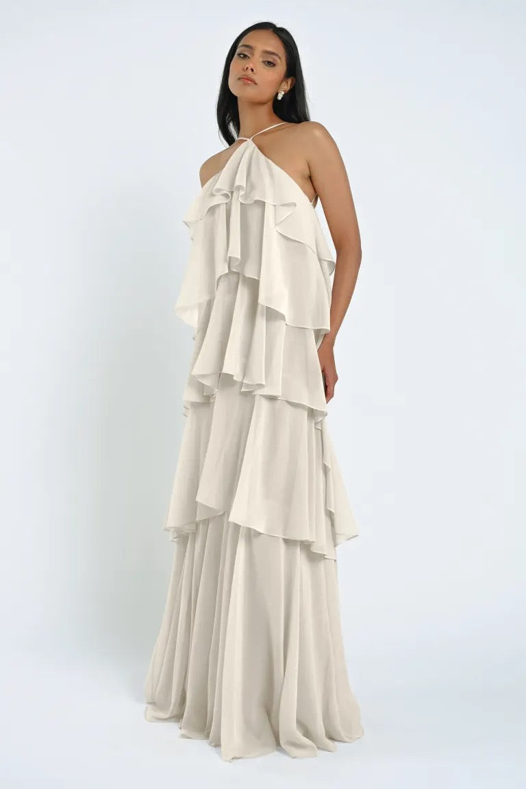 A woman stands against a plain background, wearing a floor-length, halter-neck chiffon dress featuring multiple cascading ruffles. The dress is the Farley Bridesmaid Dress by Jenny Yoo from Bergamot Bridal.