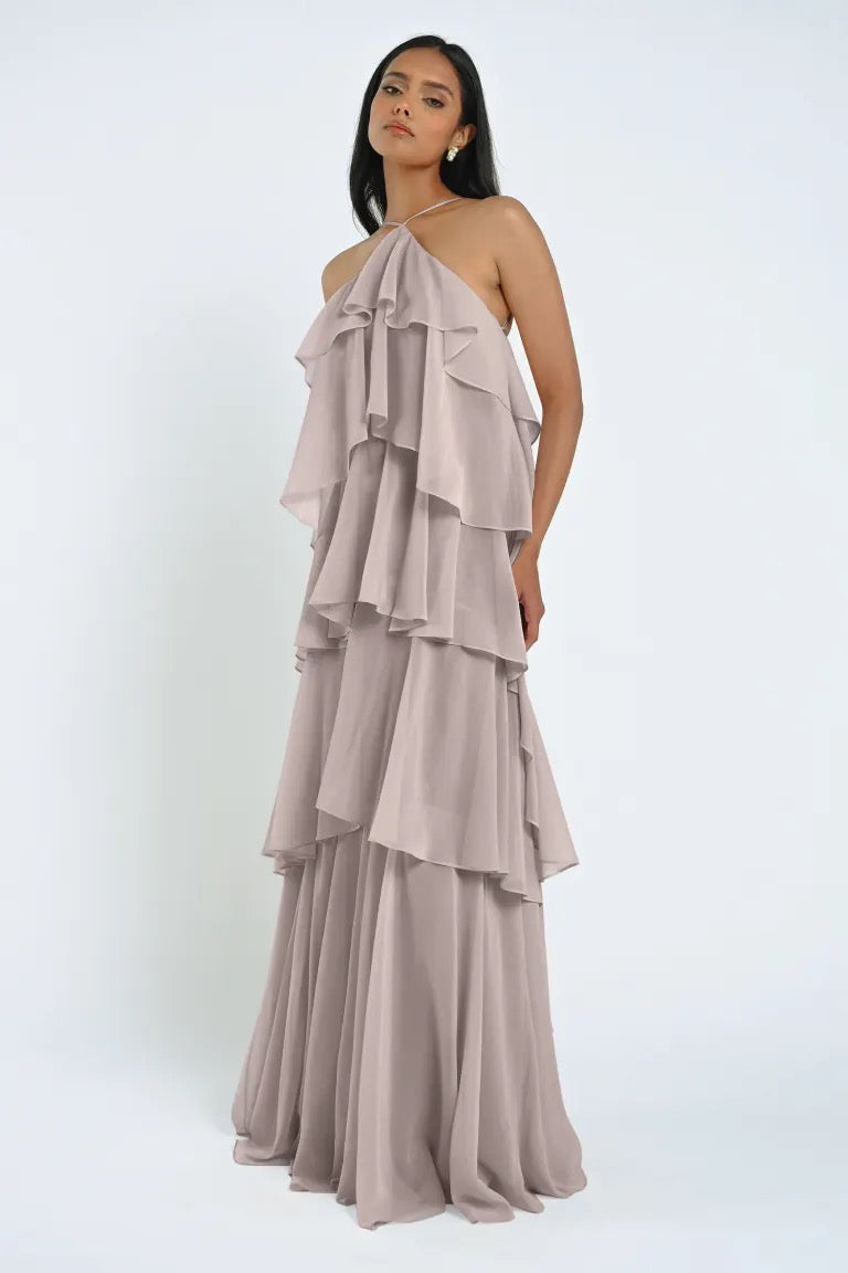 A person stands gracefully in the Farley Bridesmaid Dress by Jenny Yoo, a grey, tiered chiffon gown featuring a halter neckline, against a plain background, effortlessly showcasing this stunning creation from Bergamot Bridal.