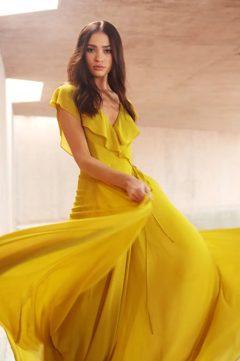 A woman in a flowing yellow chiffon Faye - Bridesmaid Dress by Jenny Yoo stands in a room with wooden architecture from Bergamot Bridal.