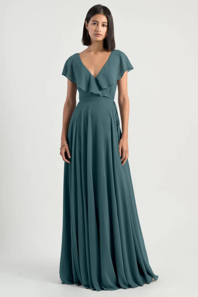 A woman in a floor-length, teal Faye bridesmaid dress by Jenny Yoo from Bergamot Bridal with a V-neck and short flutter sleeves stands against a plain background, showcasing her romantic attire with its adjustable waist for the perfect fit.