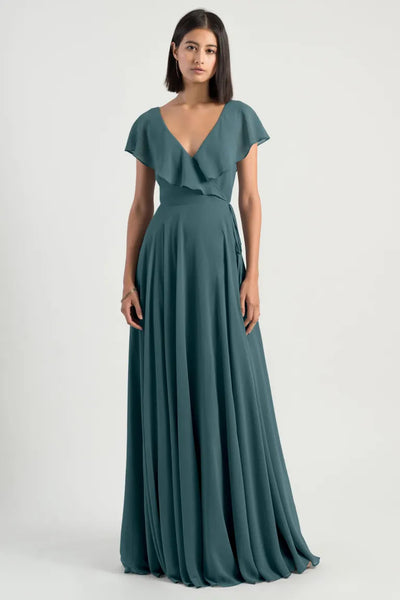 A woman in a floor-length, teal Faye bridesmaid dress by Jenny Yoo from Bergamot Bridal with a V-neck and short flutter sleeves stands against a plain background, showcasing her romantic attire with its adjustable waist for the perfect fit.