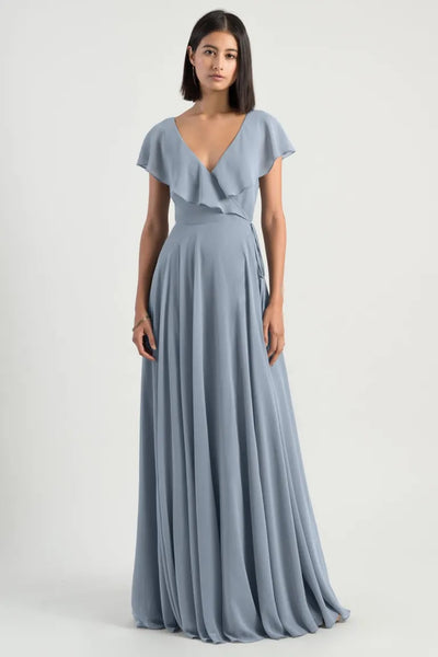 A woman in a Faye bridesmaid dress by Jenny Yoo from Bergamot Bridal, which is a grey v-neck evening gown with flutter sleeves, stands against a white background.