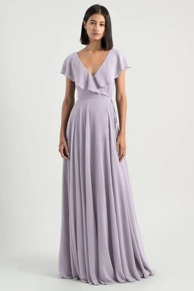 Woman posing in a full-length, lavender chiffon Faye bridesmaid dress by Jenny Yoo with a v-neckline and flutter sleeves from Bergamot Bridal.
