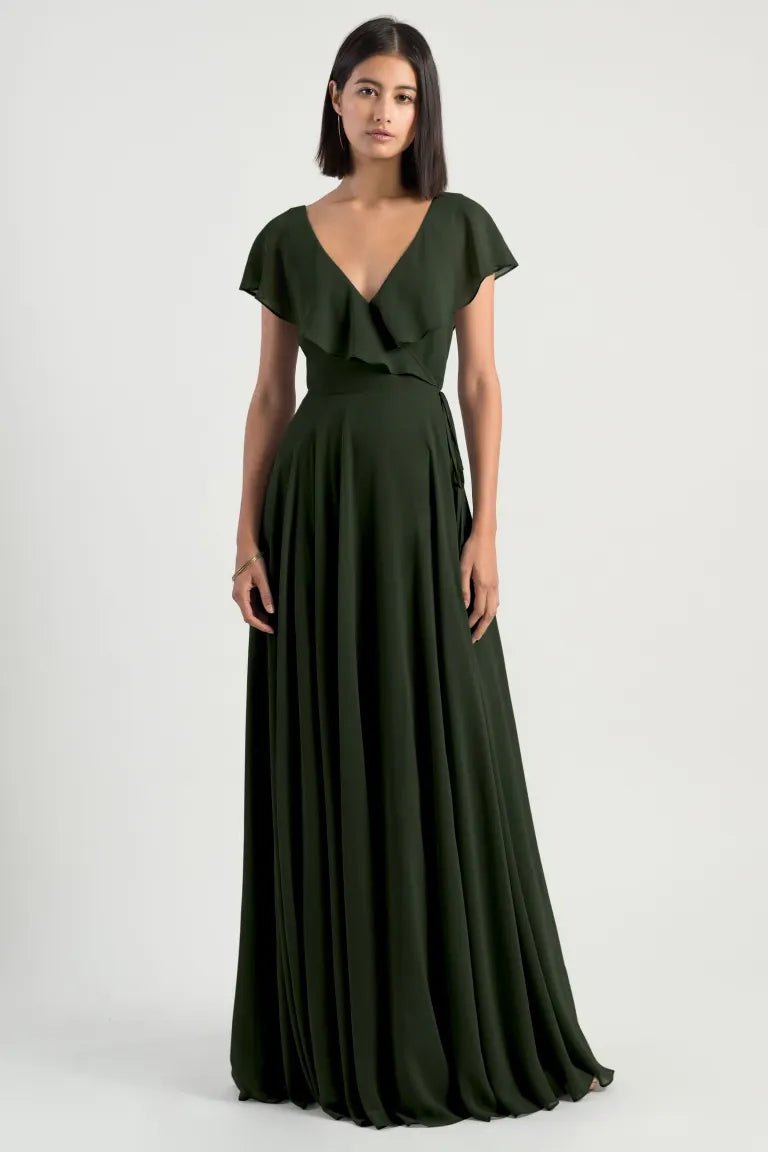 A model wearing a floor-length, dark green Faye - Bridesmaid Dress by Jenny Yoo from Bergamot Bridal, featuring a deep V-neckline and short sleeves, standing against a plain background.