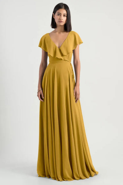A woman in an elegant mustard yellow chiffon wrap dress with a plunging neckline, draped details, and flutter sleeve by Faye - Bridesmaid Dress from Jenny Yoo at Bergamot Bridal.