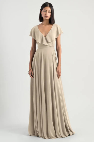 A woman standing against a neutral background, wearing an elegant beige chiffon wrap dress with flutter sleeves and a v-neckline, the Faye Bridesmaid Dress by Jenny Yoo from Bergamot Bridal.