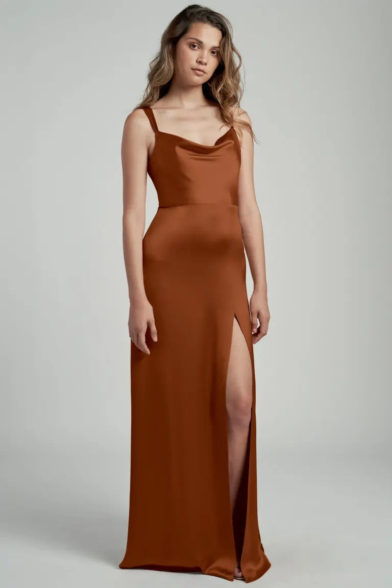 Woman posing in a Gina bridesmaid dress by Jenny Yoo from Bergamot Bridal, featuring a brown satin fabric, cowl neckline, and thigh-high slit.