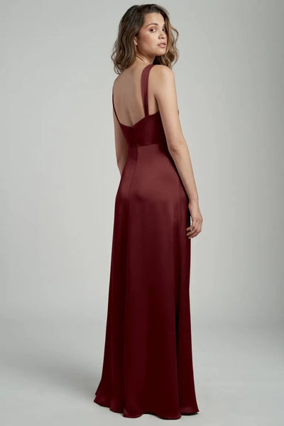 Woman in an elegant burgundy satin Gina - Bridesmaid Dress by Jenny Yoo with a cowl neckline, viewed from the back from Bergamot Bridal.