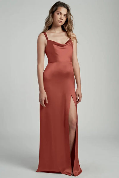 A woman in an elegant Gina bridesmaid dress by Jenny Yoo in terracotta satin with a thigh-high slit and a cowl neckline, posing against a neutral background.