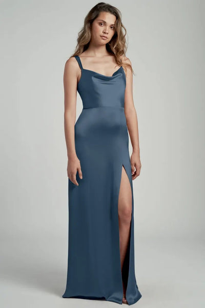 A woman in a sleek blue satin Gina bridesmaid dress by Jenny Yoo with a cowl neckline and a side slit posing against a neutral background. (Brand Name: Bergamot Bridal)