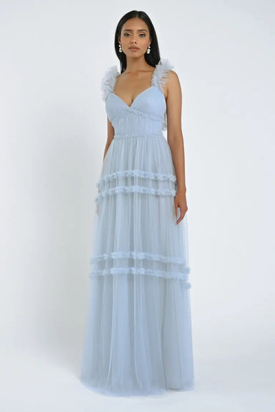 A woman wearing the Graham Bridesmaid Dress by Jenny Yoo from Bergamot Bridal, a light blue, floor-length romantic dress with tulle ruffles and tiered layers, poses against a plain white background.