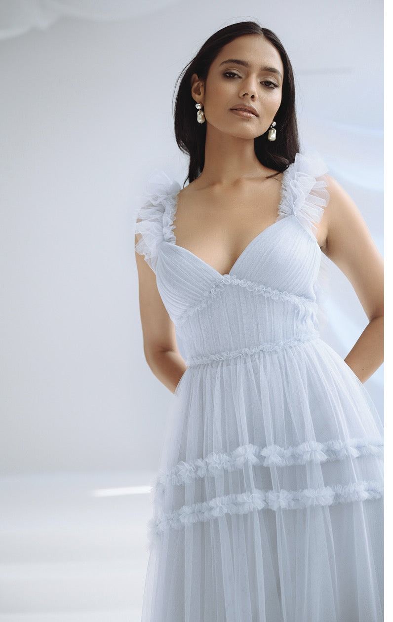 A woman with long dark hair wears a light blue, sleeveless Graham Bridesmaid Dress by Jenny Yoo from Bergamot Bridal, adorned with ruffles that create a romantic effect. She stands with one hand on her hip against a soft, light-colored background.