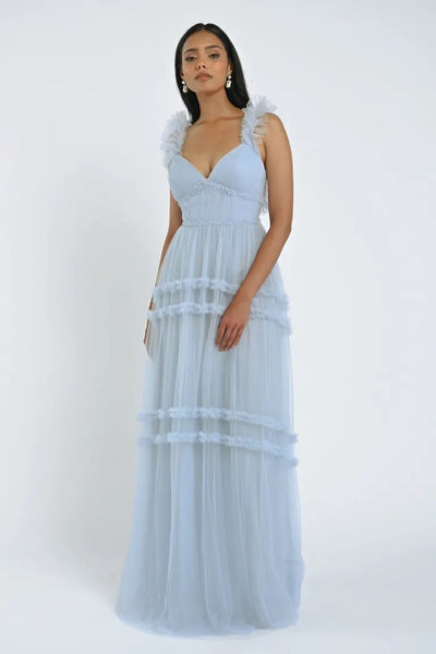 A woman stands wearing the light blue sleeveless Graham Bridesmaid Dress by Jenny Yoo, available at Bergamot Bridal, featuring ruffled shoulder straps and a layered skirt that create a romantic effect. She holds a small clutch in her right hand and looks directly forward.