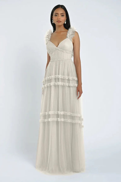 A woman in a white sleeveless Graham Bridesmaid Dress by Jenny Yoo, offered by Bergamot Bridal, with ruffled shoulders and tiered tulle layers stands against a plain background, creating a romantic effect.