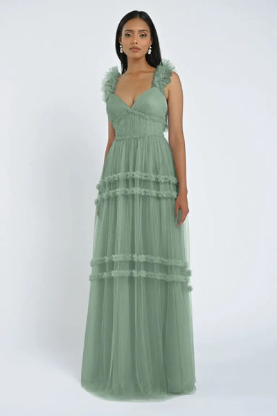 A woman stands wearing the Graham Bridesmaid Dress by Jenny Yoo, a long, romantic sage green dress featuring tiered tulle and ruffled sleeves. She has straight hair and is facing the camera against a plain white background. This elegant piece is part of Bergamot Bridal's collection.