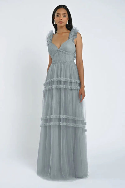 A person stands wearing a light blue, sleeveless Graham Bridesmaid Dress by Jenny Yoo from Bergamot Bridal, featuring ruffled details and a V-neckline, creating a romantic effect against the plain white background.