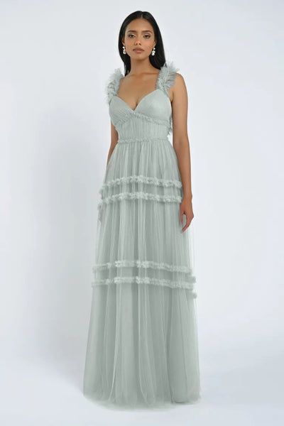 A woman stands wearing the Graham Bridesmaid Dress by Jenny Yoo from Bergamot Bridal, a floor-length light grey dress with romantic ruffled sleeves and tiered layers, a true showstopper.