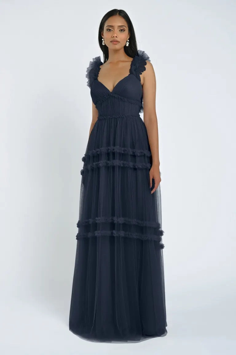 A woman stands wearing the Graham Bridesmaid Dress by Jenny Yoo from Bergamot Bridal, a floor-length, dark-colored gown with ruffled shoulder straps and tiered layers, creating a romantic effect.