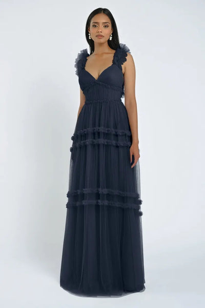 A woman stands wearing the Graham Bridesmaid Dress by Jenny Yoo from Bergamot Bridal, a floor-length, dark-colored gown with ruffled shoulder straps and tiered layers, creating a romantic effect.