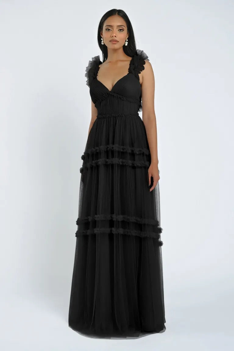 Description: Woman wearing the Graham Bridesmaid Dress by Jenny Yoo, a long, black, sleeveless showstopper with tulle ruffle details from Bergamot Bridal, standing against a plain white background.