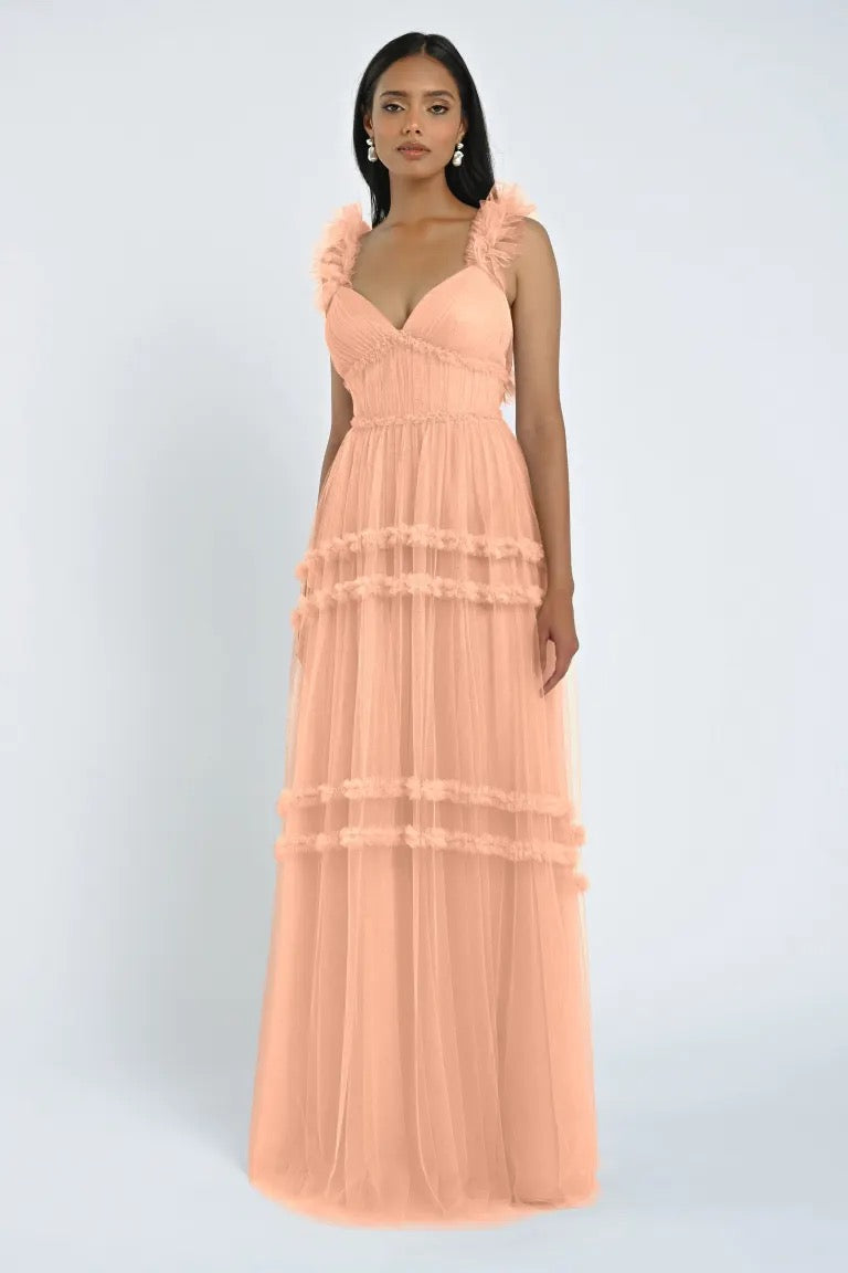 A woman stands wearing the Graham Bridesmaid Dress by Jenny Yoo from Bergamot Bridal, a long peach-colored gown with ruffled straps and layered tulle details that showcase a truly romantic look.