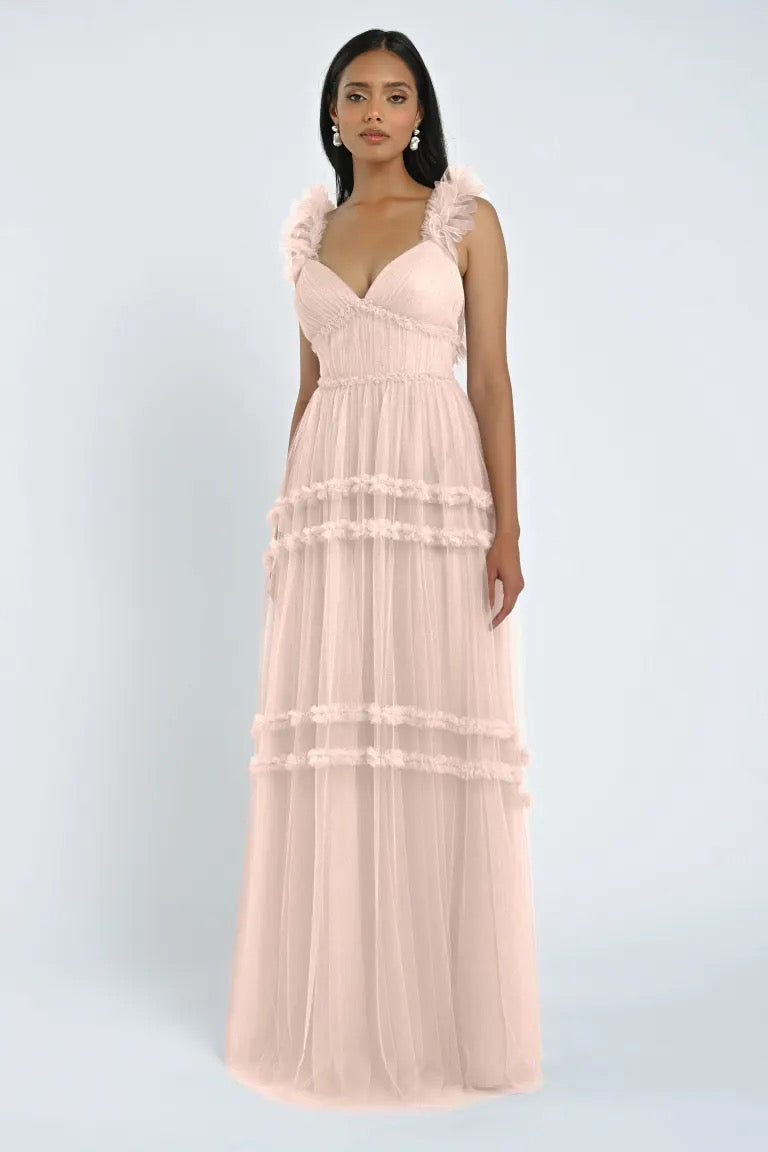 Woman wearing the Graham Bridesmaid Dress by Jenny Yoo from Bergamot Bridal, a light pink sleeveless dress featuring romantic tulle ruffles and a tiered skirt, posing against a light background.