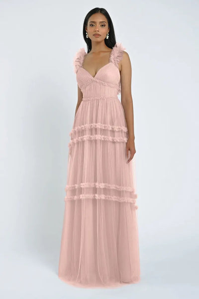 A woman is wearing a romantic Graham Bridesmaid Dress by Jenny Yoo from Bergamot Bridal, a long pink gown with tulle ruffles and tiered tulle layers. She stands against a light-colored background.