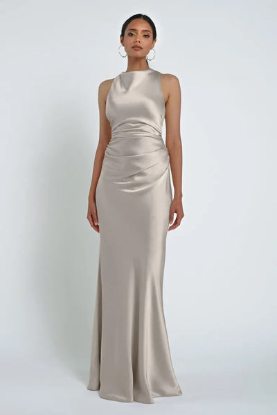 Person standing wearing a long, sleeveless Grant Bridesmaid Dress by Jenny Yoo from Bergamot Bridal, made from luxe satin fabric with a high neckline against a plain white background.