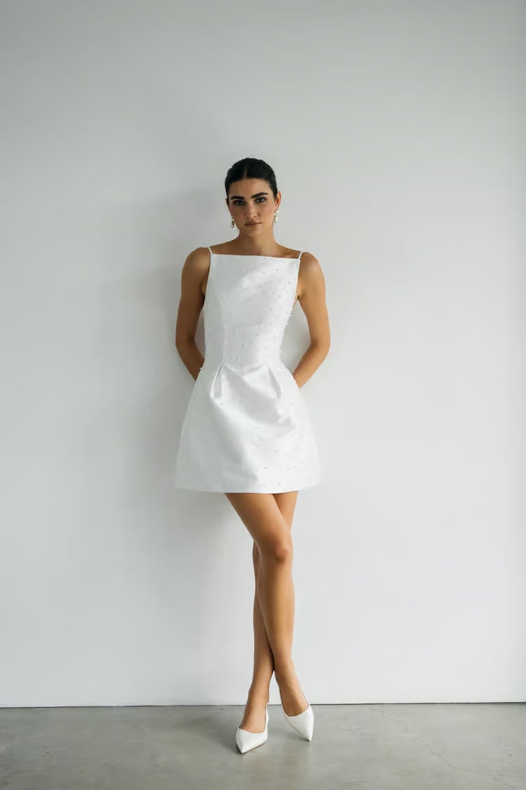 A woman stands against a white wall wearing the sleeveless, short Hannah - Jenny Yoo Little White Dress by Bergamot Bridal with white pointed heels—a chic bridal look.