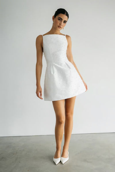 A woman stands against a white wall in the Hannah - Jenny Yoo Little White Dress by Bergamot Bridal, a sleeveless, above-the-knee style with subtle embellishments—ideal for a bridal occasion—paired with white pointed-toe heels.