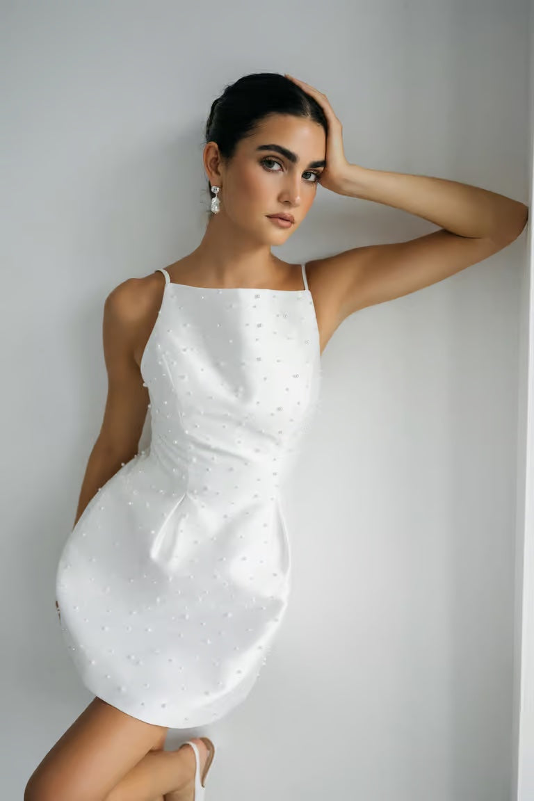 A woman in the sleeveless, pearl-embellished Hannah - Jenny Yoo Little White Dress by Bergamot Bridal leans against a white wall, resting her left arm on her head and gazing at the camera—a lovely choice for bridal occasions.