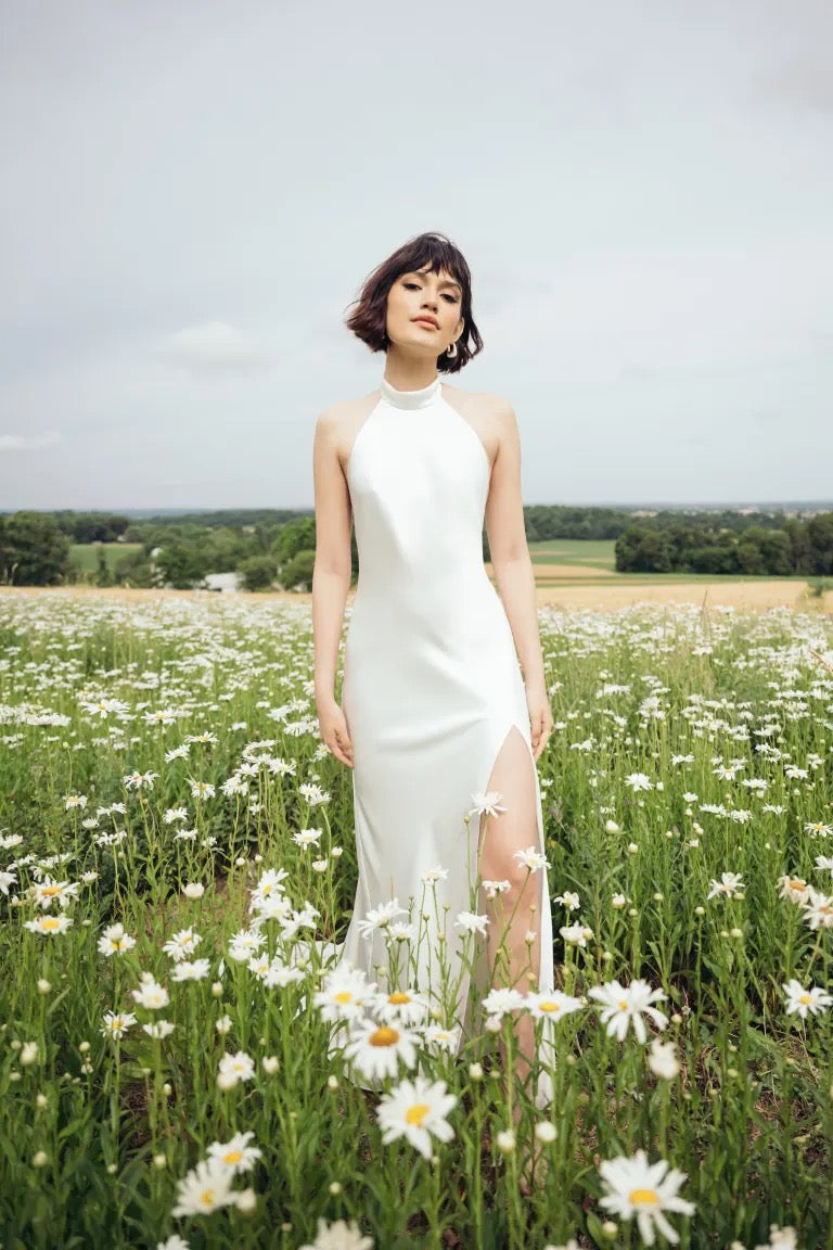 A person in a Harlyn Wedding Dress by Jenny Yoo from Bergamot Bridal stands gracefully in a field of daisies under a cloudy sky.