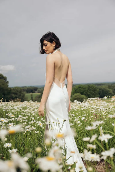 A woman in a backless Harlyn Wedding Dress by Jenny Yoo from Bergamot Bridal stands in a field of daisies, the luxe satin fabric shimmering as she looks over her shoulder.