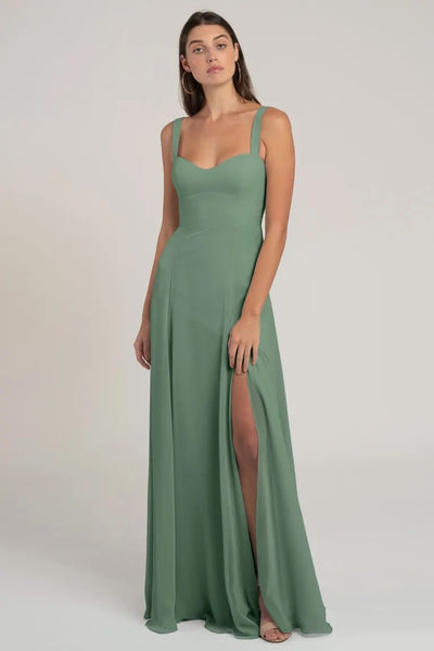 A woman standing in a studio wearing an elegant green chiffon Harris bridesmaid dress by Jenny Yoo with a sweetheart neckline and a thigh-high slit.