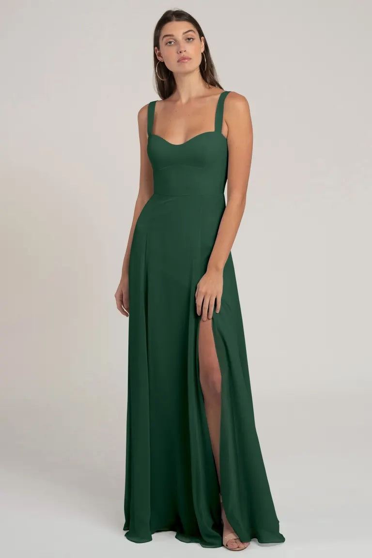 A woman in an elegant green Harris - Bridesmaid Dress by Jenny Yoo with a flattering silhouette and a high slit standing against a neutral background.