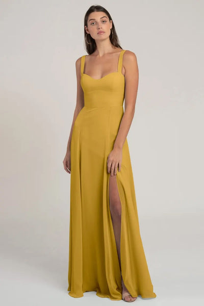 A woman stands posing in a long, mustard yellow chiffon evening dress with a bombshell neckline and a thigh-high slit. - Harris Bridesmaid Dress by Jenny Yoo from Bergamot Bridal.