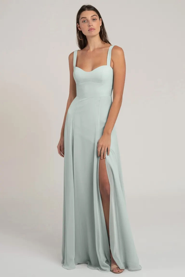 A woman stands wearing a Harris - Bridesmaid Dress by Jenny Yoo in pale blue chiffon with a sweetheart neckline and a thigh-high slit from Bergamot Bridal.