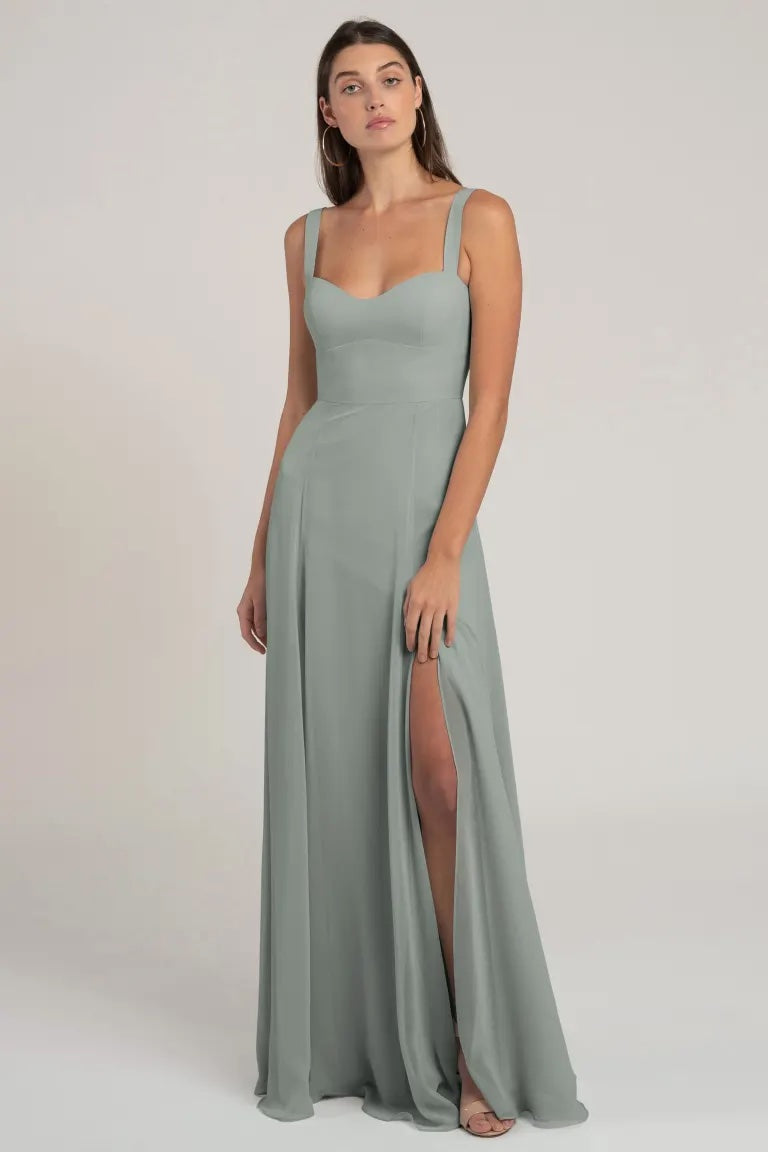 A woman in an elegant, grey sleeveless chiffon bridesmaid dress with a high slit and a flattering silhouette, standing against a neutral background. The dress is the Harris Bridesmaid Dress by Jenny Yoo from Bergamot Bridal.