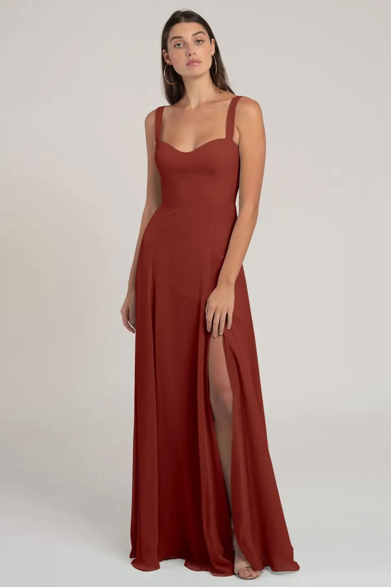 Woman posing in an elegant rust-colored Harris bridesmaid dress by Jenny Yoo with a flattering silhouette and a slit, from Bergamot Bridal.