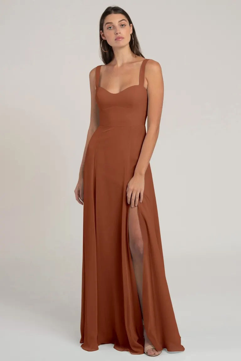 A woman posing in an elegant, sleeveless, brown chiffon Harris bridesmaid dress by Jenny Yoo with a high slit and a flattering silhouette from Bergamot Bridal.