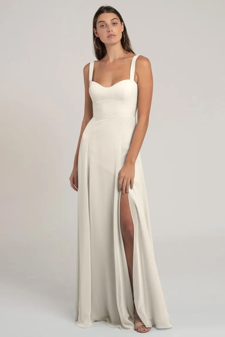 A woman standing in a flattering silhouette, wearing a simple and elegant white gown with a slit up the side - Harris Bridesmaid Dress by Jenny Yoo from Bergamot Bridal.