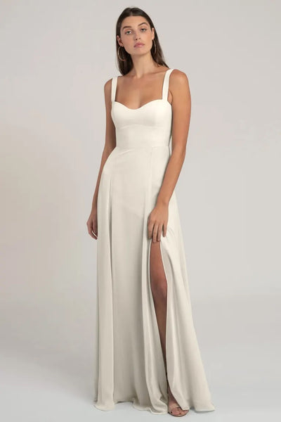 A woman standing in a flattering silhouette, wearing a simple and elegant white gown with a slit up the side - Harris Bridesmaid Dress by Jenny Yoo from Bergamot Bridal.