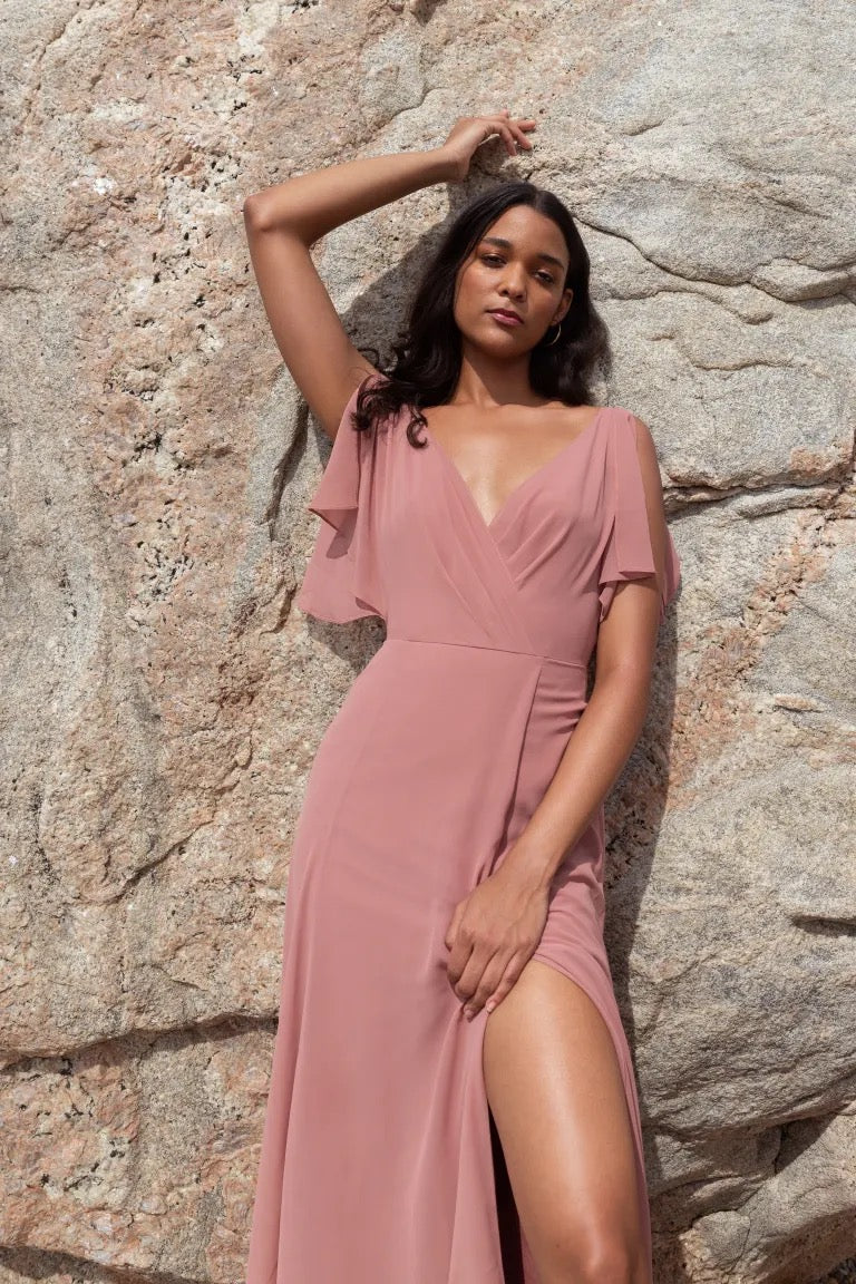 A woman in a beautiful Hayes - Bridesmaid Dress by Jenny Yoo with flutter sleeves posing against a rocky background from Bergamot Bridal.