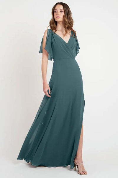 A woman in a floor-length, teal Hayes - Bridesmaid Dress by Jenny Yoo from Bergamot Bridal, featuring flutter sleeves and a V-neckline, stands against a plain white background. She is wearing strappy high-heeled sandals. The beautiful dress is showcased as a store sample size 24.