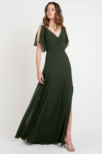 Woman wearing the stunning Hayes - Bridesmaid Dress by Jenny Yoo in a deep green hue, showcasing its elegant flutter sleeves and V-neckline, as she poses against a simple white background. The dress is by Bergamot Bridal.